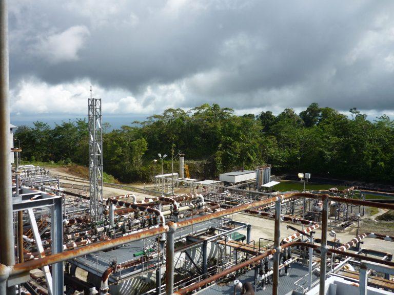 Industrial facility with complex piping, surrounded by greenery and a cloudy sky in the background.