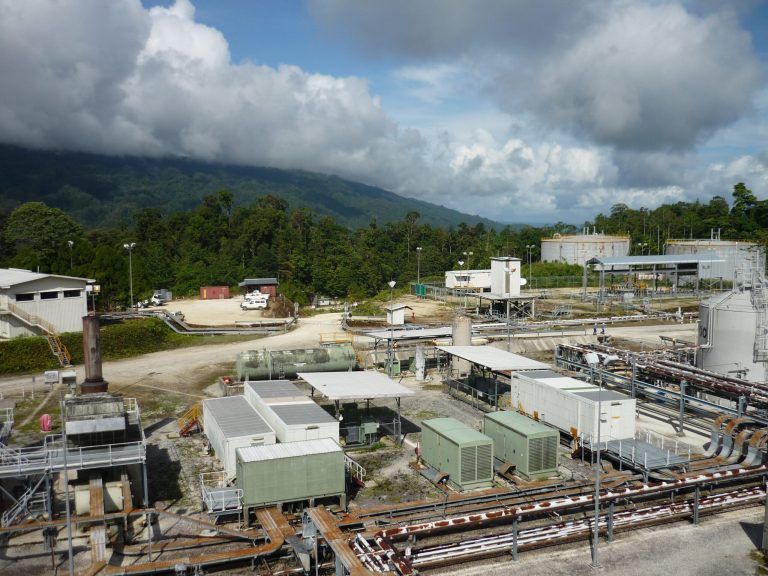 Industrial facility surrounded by lush greenery and mountains under a cloudy sky.