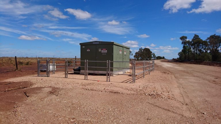 Fenced green utility box on a dirt road, with a clear sky and sparse vegetation in the background.