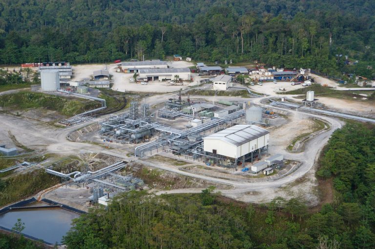 Aerial view of an industrial facility with multiple buildings and pipelines surrounded by forest.