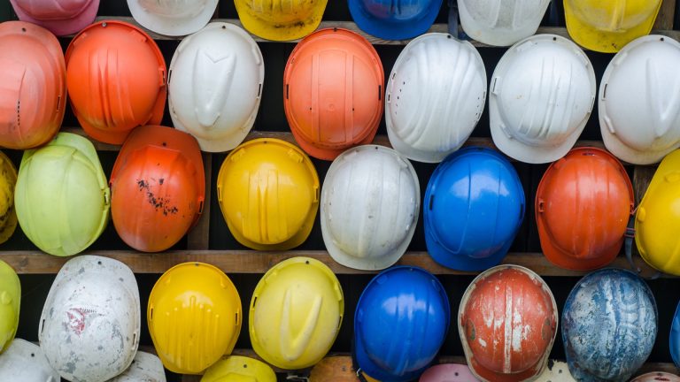 A collection of construction hard hats in various colors, including yellow, orange, blue, and white, displayed in rows.