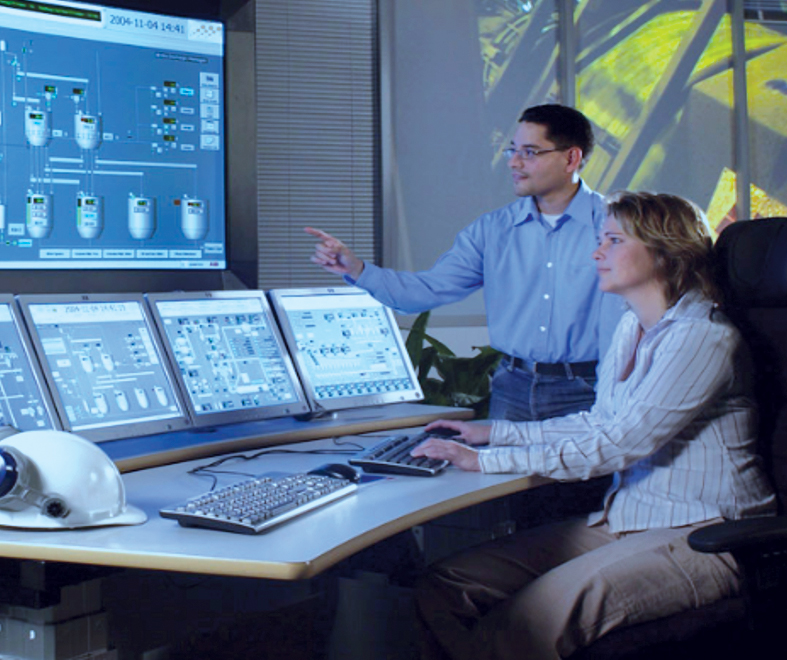 Two people working at a control room desk with multiple monitors showing technical schematics.