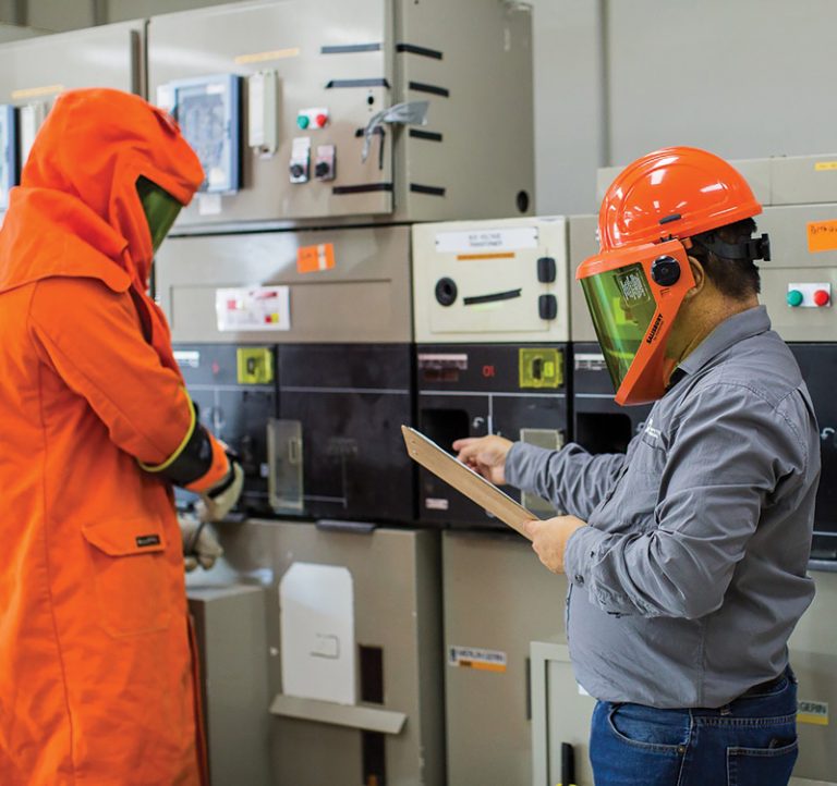 Two workers in protective gear operating electrical panels in an industrial setting.