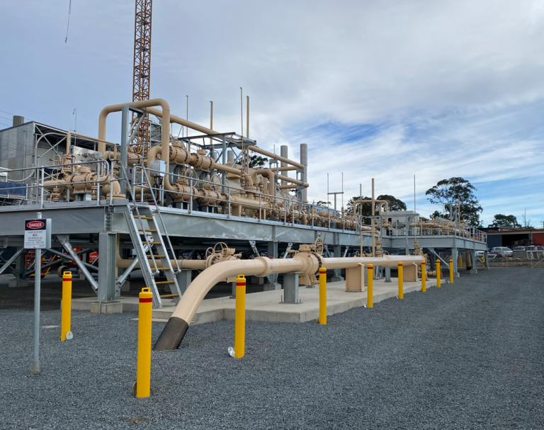 A gas processing facility with beige piping and equipment, surrounded by gravel and safety bollards under a cloudy sky.