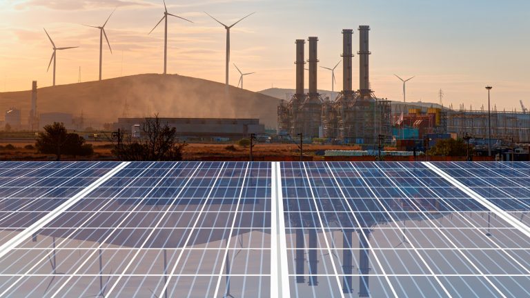 A solar farm with panels in the foreground, wind turbines, and an industrial facility with smoke in the background at sunset.