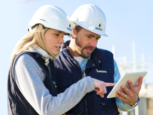 Two Verbrec employees in safety helmets and vests reviewing information on a tablet at a worksite.