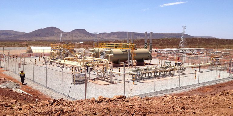 Industrial facility in a desert landscape with fenced equipment and pipelines, set against distant mountains.